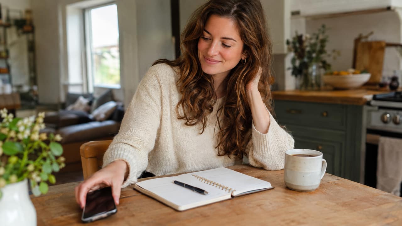 Woman sitting on her living room sofa having a relaxed phone conversation with a notebook open beside her