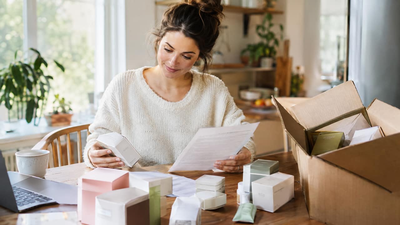 A person at a kitchen table sorting boxes of unopened product while reviewing paperwork on a laptop
